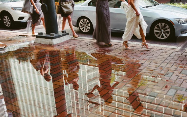 Bride and bridal party legs reflect in rain puddle, leave hotel through rain soaked streets in Houston, Texas.