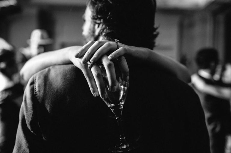 During wedding reception a woman's arms drape around mans shoulders with champagne glass