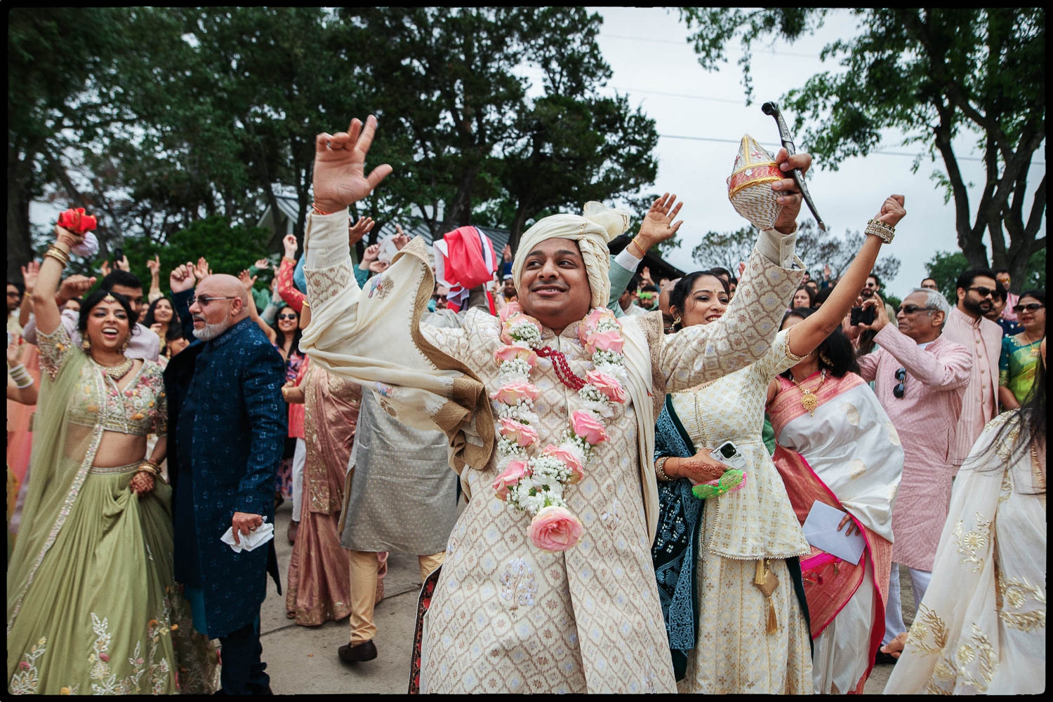 a man in a white dress with a flower garland and a hat with his hands up during the baraat